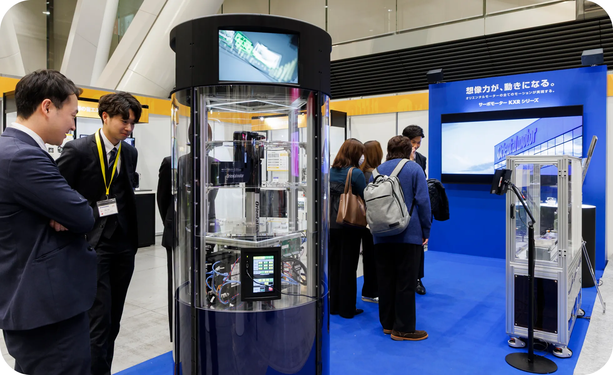 円柱形のガラスケースに収められた最新機器を興味深く見つめる来場者たち。