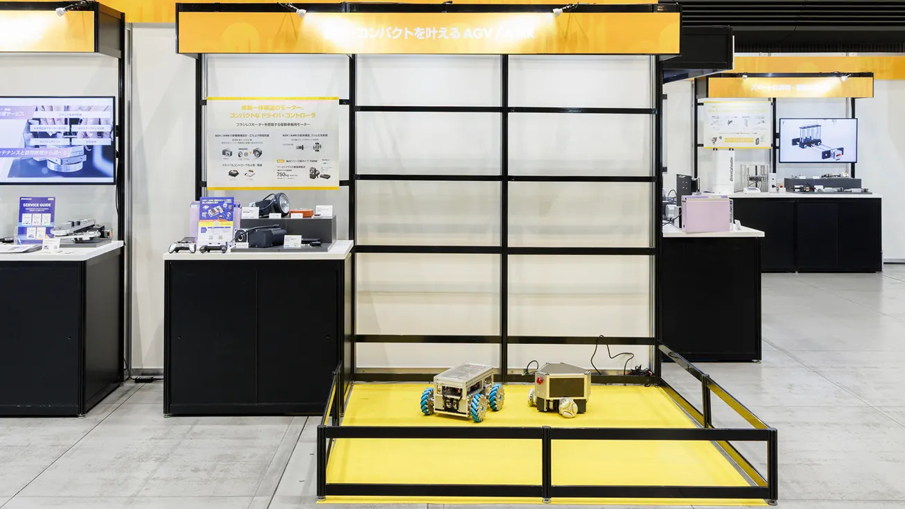 「コンパクトを叶えるAGV/AMR」をテーマとした展示ブース。中央のデモスペースでは、メカナムホイールを搭載した小型の搬送ロボットが2台展示されている。